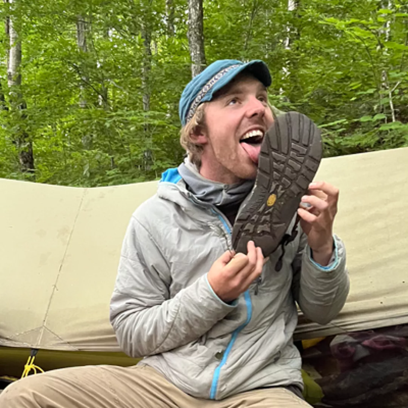 The image shows a man outdoors, seemingly camping. He is holding a shoe and licking the sole with a look of amusement. He is wearing a hat and a jacket, suggesting a cool environment. The background includes trees and a tent, indicating a natural setting.